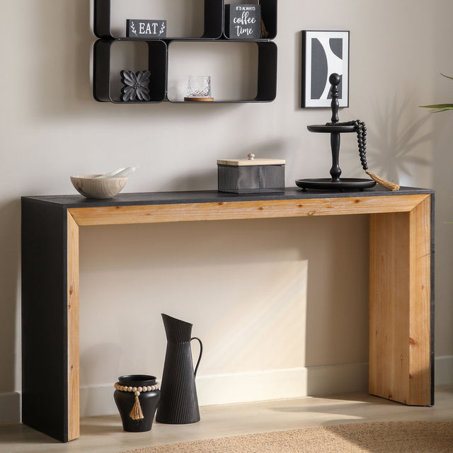 A rectangular wooden console table with black finish on top and natural wood legs, featuring a mix of black and natural wood colors. The table is positioned against a wall with decorative items on it, such as a vase with flowers, a bowl, and framed pictures.