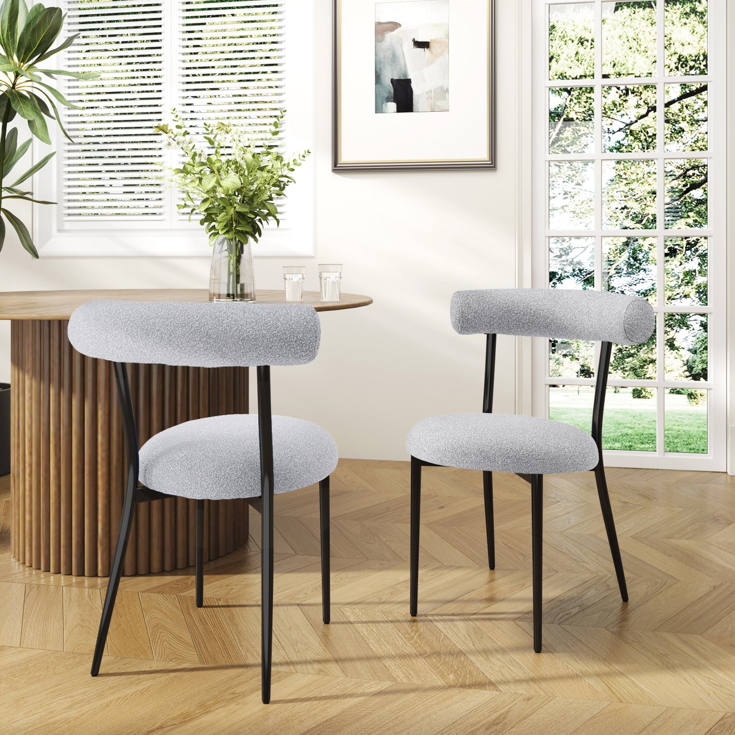 Two modern upholstered side chairs with black legs and gray fabric seats and backrests, set against a backdrop of a well-lit room with a wooden floor and a round wooden table in the foreground.