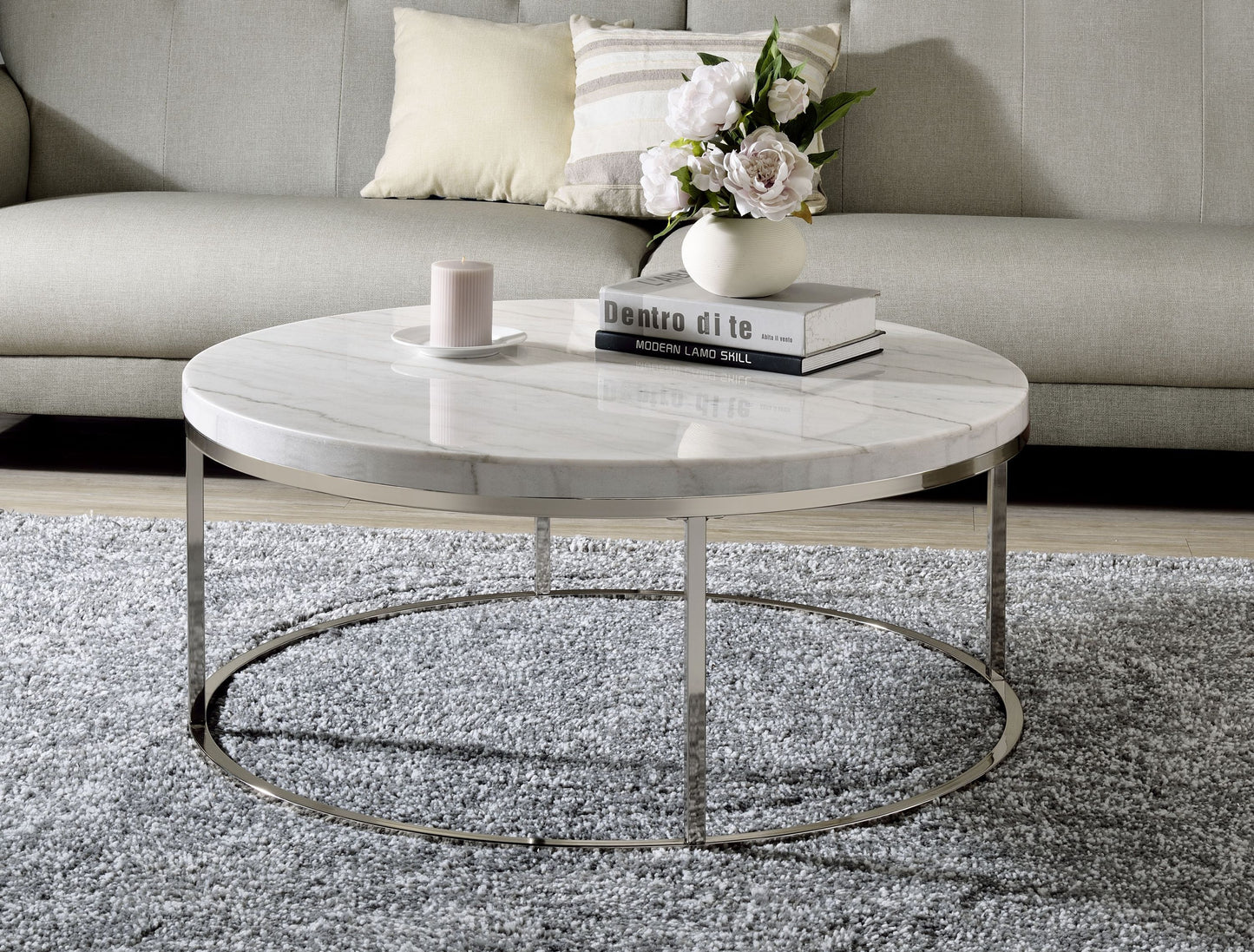 A round coffee table with a marble top and nickel finish metal frame, placed on a grey area rug in a cozy living room setting with a couch, pillows, and a vase with flowers in the background.