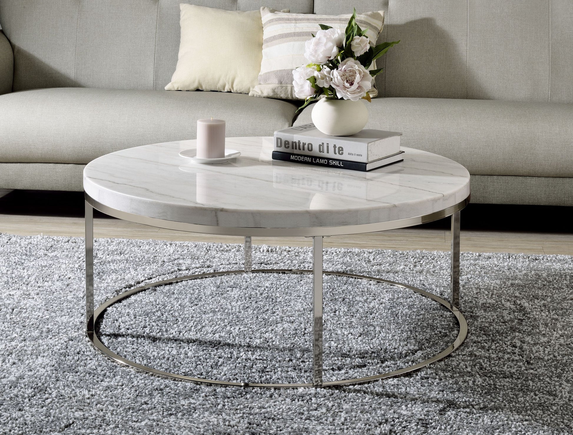 A round coffee table with a marble top and nickel finish metal frame, placed on a grey area rug in a cozy living room setting with a couch, pillows, and a vase with flowers in the background.