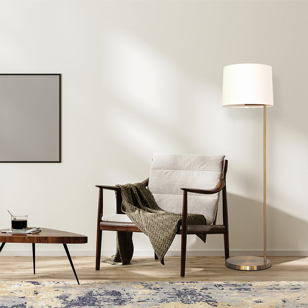 An elegant brass floor lamp with a drum-shaped linen shade, placed in a room with a chair and a small table.